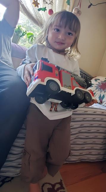 A young child with blonde hair and a white long-sleeve shirt with brown pants holds up a large red and white toy fire truck, smiling at the camera.
