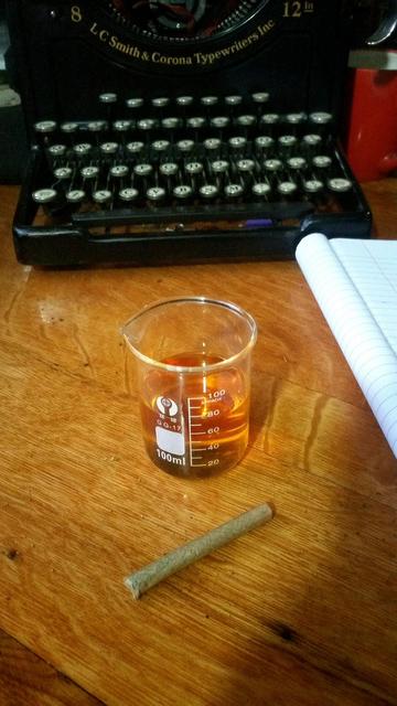 a 100ml beaker 3/4 full of bottom/mid tier whisky and a spliff next to a notebook before a typewriter all on a wooden table