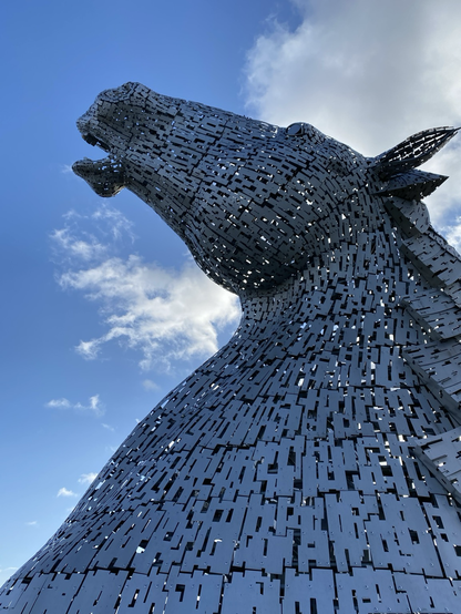 I took this photo a few years ago. It shows one of The Kelpies, two 30 metre monumental high horses heads sculptures by artist Andy Scott. This is the highest equine sculpture in the world. Constructed of stainless steel shards, this powerful horse’s head is looking upwards into the sky with light permeating the metal structure. The sheer scale and effect of the light are stunning, dominating the surrounding area. All against a blue sky with a wispy cloud or two. This commemorates industrial heritage and kelpie folklore. A must-see if you are ever in Scotland.