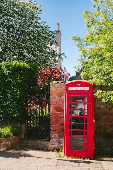 A classic old red British telephone box on a paved street, a red stone-brick wallbehind it with a black wrought iron gate in the wall next to a hedgerow, a variety of trees and bushes behind the wall, and the edge of a house with a tall chimney just visible against a pale blue sky.