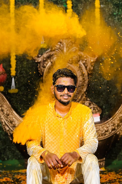 An Indian man in loose white clothing and shades sitting on a floor strewn with orange petals. There's an explosion of yellow pigment powder around him, staining his shirt bright yellow and his trousers a paler pastel. There's an ornate golden sculpture behind him, and strings of yellow decoration hanging down from above.