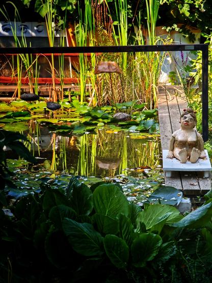 An einem recht zugewachsenen Gartenteich sitzt auf einem hölzernen Steg die Tonfigur einer Frau. (Figur von Erwin Schwentner)