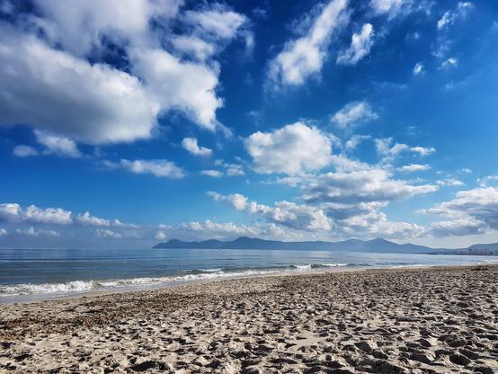 A wide-angle, low-view shot of a sandy beach stretching towards a calm sea under a vibrant blue sky dotted with fluffy white clouds. In the distance, a long, low mountain range is visible on the horizon, separated from the beach by the ocean. The foreground is dominated by the textured, light-brown sand.