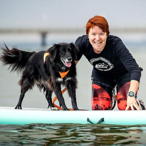 Ein schwarzer Hund auf einem SUP auf der Ostsee. Ich knie neben ihm und lache in die Kamera.