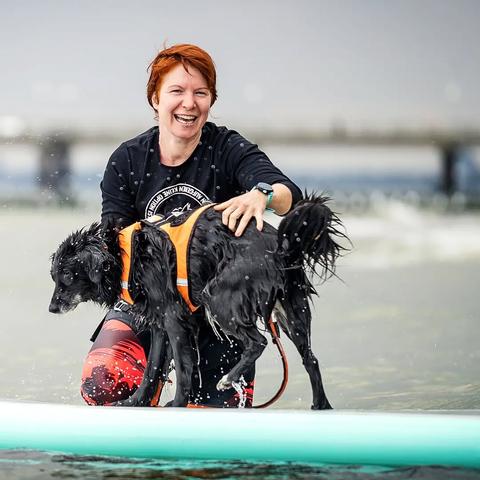 Ein schwarzer, nasser Hund auf einem SUP auf der Ostsee. Ich knie hinter ihm und lache in die Kamera.