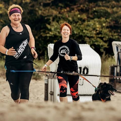 Zwei Frauen in Laufkleidung rennen mit zwei Hunden im Zuggeschirr einen Strand entlang auf die Kamera zu. Im Hintergrund sieht man Strandkörbe.