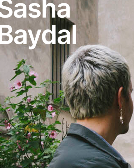 There is the picture of Sasha's head from the back. They are wearing an earring on the right ear. On the background there is a begue wall and green bushes. The name of the speaker is written in white letters "Sasha Baydal".
