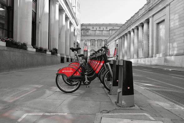A partial colour photograph of some rental bikes in london. The picture is black and white, but the red of the bikes advertising is visible. The bikes are parked in a line, and the photo shoots through the middle of all of them in a line. The surroundings are the opulent Grecian columns of the Square Mile around the City of London.