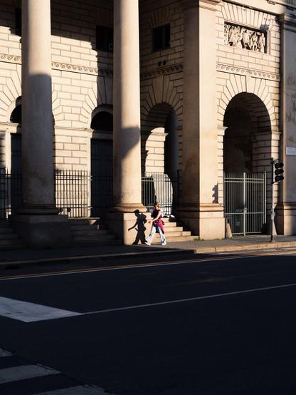 A woman walks past an ornate building's facade, partly lit by the low sunlight of sunset. She is illuminated and she projects her shadow on the base of a large column.