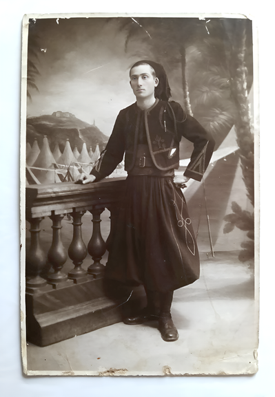 Un homme se tien debout devant un décor orientaliste, il pose pour la photo. Il porte son uniforme de zouave d'apparat, avec le pantalon bouffant typique. La photo est en noir et blanc.