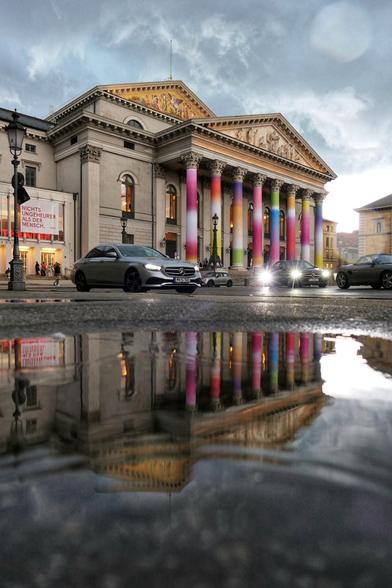 Das Bild zeigt ein großes, klassisch gestaltetes Gebäude mit einer auffälligen Fassade. Die Vorderseite des Gebäudes ist von mehreren Säulen geprägt, die in kräftigen Farben gestrichen (oder mit diesen Farben umhüllt) sind – darunter Rosa, Lila, Grün und Gelb. Über den Säulen befindet sich ein dreieckiges Giebelfeld mit einem kunstvollen Relief, das dem Bauwerk einen historischen Charakter verleiht. Links daneben, auf einem Gebäude, das laut Leuchtbuchstaben EIN Theater ist, hängt ein Banner mit der Aufschrift „NICHTS UNGEHEURER ALS DER MENSCH.“ Vor dem Gebäude fahren mehrere Autos oder sind geparkt , darunter ein silberner Mercedes. Eine Pfütze im Vordergrund reflektiert sowohl das Gebäude als auch die Fahrzeuge, was dem Bild eine zusätzliche räumliche Tiefe verleiht. Der Himmel ist grau und wolkenverhangen, was auf regnerisches oder trübes Wetter hindeutet.