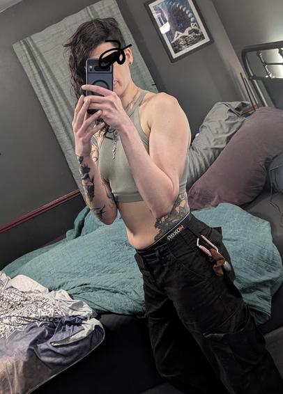 This enby taking a mirror selfie in front of their bed. They're wearing black cargo pants with pockets spacious enough to hide secrets. A pink carabiner hangs from their belt loop holding a set of earplugs and a brown scrunchie. The waistband of their rainbow and black woxers peeks from under the cargo pants and demarcates the tattoo of wildflowers, mushrooms and mandala along the side of their torso. The mandala sits at the curve of the small of the back clearly indicating stable hip mass. 

The soft skin is again concealed by a sage green crop top and both arms holding the camera phone in front of their face. The jellyshroom tendrils and the owl portal are the visible parts of their right arm, while their left arm is just smooth skin, toned muscle and creamy squish layered over. Their hair is messy and falling to the far side. They're wearing three pendants. The non-binary, amethyst and salamander in increasingly sharp Vs down the center of their abdomen lightly indicating some of the hidden cleavage being squished away by the arms. They seem to be trying to focus and have inadvertently left a bit of their tongue hanging out. It's used for balancing fine motor movements when phones don't want to cooperate.