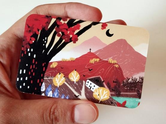 Le verso de la carte. Ca représente un paysage montagneux, en teintes rouges ocres brunes et doré... Avec une ferme comtoise sur un mont, et une lune noire perchée dans le ciel jaune. A gauche il y a un arbre au tronc noir et aux feuilles rouges.