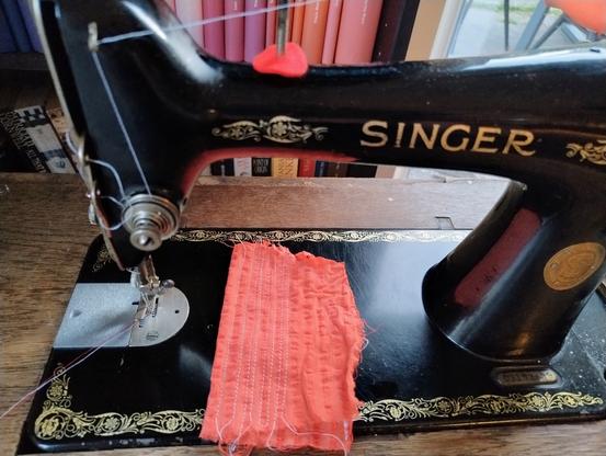 An old Singer 66 sewing machine in working condition. The machine has the classic filigree decal, but the standard text on top of the arm is missing.


 It's threaded with white thread, a piece of red fabric is under the arm of the machine. Some test stitching on the fabric. The machine is placed before a bookshelf.