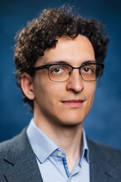 The image shows a person with curly brown hair and glasses, wearing a light blue collared shirt and a grey suit jacket. They are looking slightly upwards with a thoughtful expression. The background is a soft, blurred blue, creating a professional and serene atmosphere.