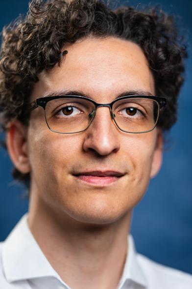 The image shows a person with curly, dark brown hair and glasses, wearing a white collared shirt. They have a gentle smile and are facing the camera against a blurred blue background, creating a focused, warm expression.
