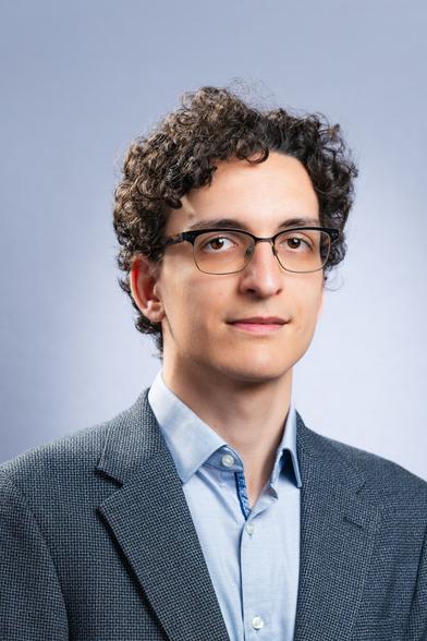 The image shows a person with curly hair and glasses, wearing a light blue collared shirt and a grey textured blazer. They have a calm, composed expression and are facing the camera with a soft blue gradient background, creating a professional and serene atmosphere.