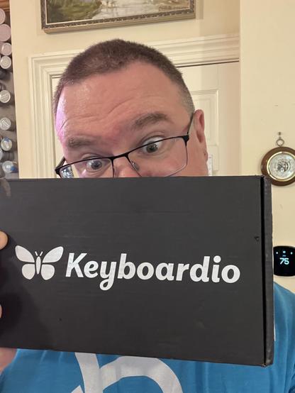 Excited-looking dude peering over the top of a black cardboard box, about a handspan in its longest dimension, labeled “Keyboardio” in white text.