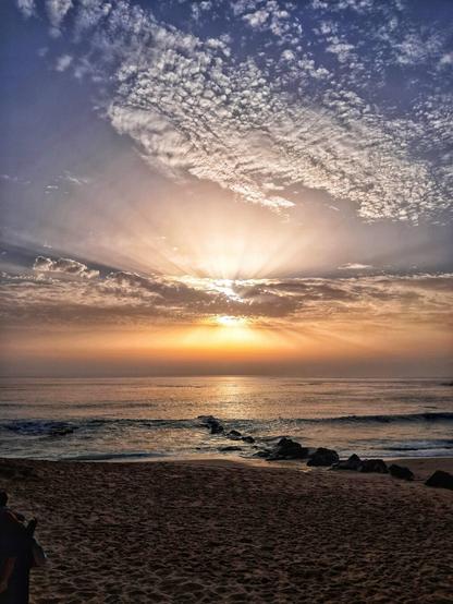 [pt] pôr do sol sobre o mar, com areia e rochas em primeiro plano. O sol brilha no céu azul, entre algumas nuvens brancas

[en] sunset over the sea, with sand and rocks in the foreground. The sun shines in the blue sky, between a few white clouds