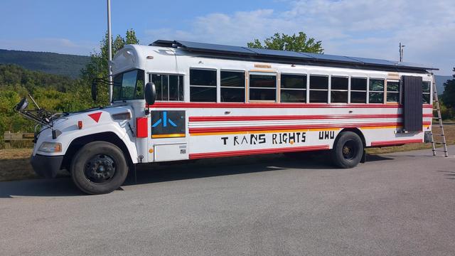 The Vulpibus parked at the side of a large tarmac area with beautiful forested hills in the background. The message on the side of the bus reads "TRANS RIGHTS UWU". The Put Together logo is also seen on the electrical box door of the driver cab. A ladder is leaning on the back of the bus and the roof has solar panels along it. The bus is an ex school bus painted red and white.
