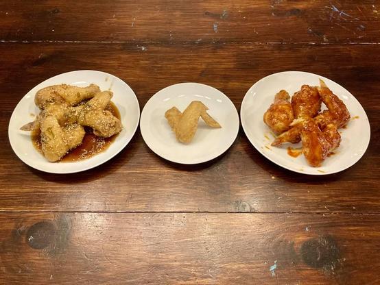 A photo of the three plates of fried chicken on a table. One has several wings and drumsticks in the Soy-Ginger sauce. Another hass a single un-sauced wing, and the third has wings and drumsticks in the Korean Chile sauce.
