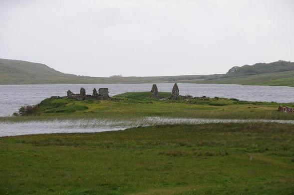 Finlaggan near Port Askaig on the Isle of Islay. The image shows a loch surrounded by low-lying land and rough grass in the foreground. A promontory extends in from the right beyond a marshy area and there are the remains of two or three ruined stone buildings on it. It is a dull day.