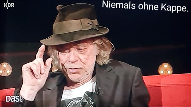 Helge Schneider tippt mit zeigefinger an hutkrempe, spruch: niemals ohne Kappe.