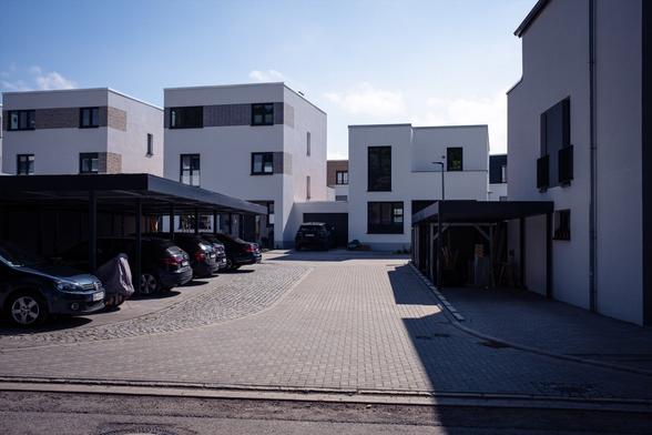 Ein modernes Wohngebiet mit mehreren weißen und grauen würfelförmigen Gebäuden mit großen Fenstern, geparkten Autos in einer gepflasterten Auffahrt und einigen schattierten Abschnitten. Der Himmel ist klar und blau. Gesamteindruck: grau.