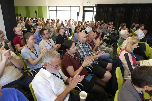 Publikum in einem Konferenzraum applaudiert während einer Veranstaltung; viele Menschen sitzen in Reihen auf grünen Stühlen.