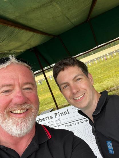 Two smiling men take a selfie on a shooting range between details. A plotting board is visible where one of them will mark the fall of shot and estimate scores for the benefit of spectators.