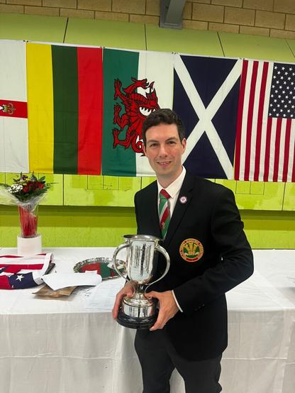 A man stands in front of a table and a row of flags holding a silver trophy. He is suited with a red and green tie and a black blazer bearing a gold, red and green badge. The badge reads "Welsh Target Shooting Federation" and is embroidered with the Prince of Wales Feathers.