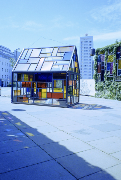 Farbfotografie. Ein kleines Glashaus aus bunten und durchsichtigen Wänden und Dachgiebel auf einem hellen Betonweg. Das Haus wirft einen bunten Schatten nach rechts. Rechts davon im Hintergrund eine bunte, mit Pflanzen umwachsene Glaswand. Im Hintergrund Wohnhäuser.