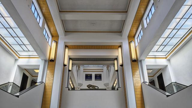 Bright mostly white high hallway, with some yellow lines and rectangular window, with staircases on either side of the balustrade in the first floor.