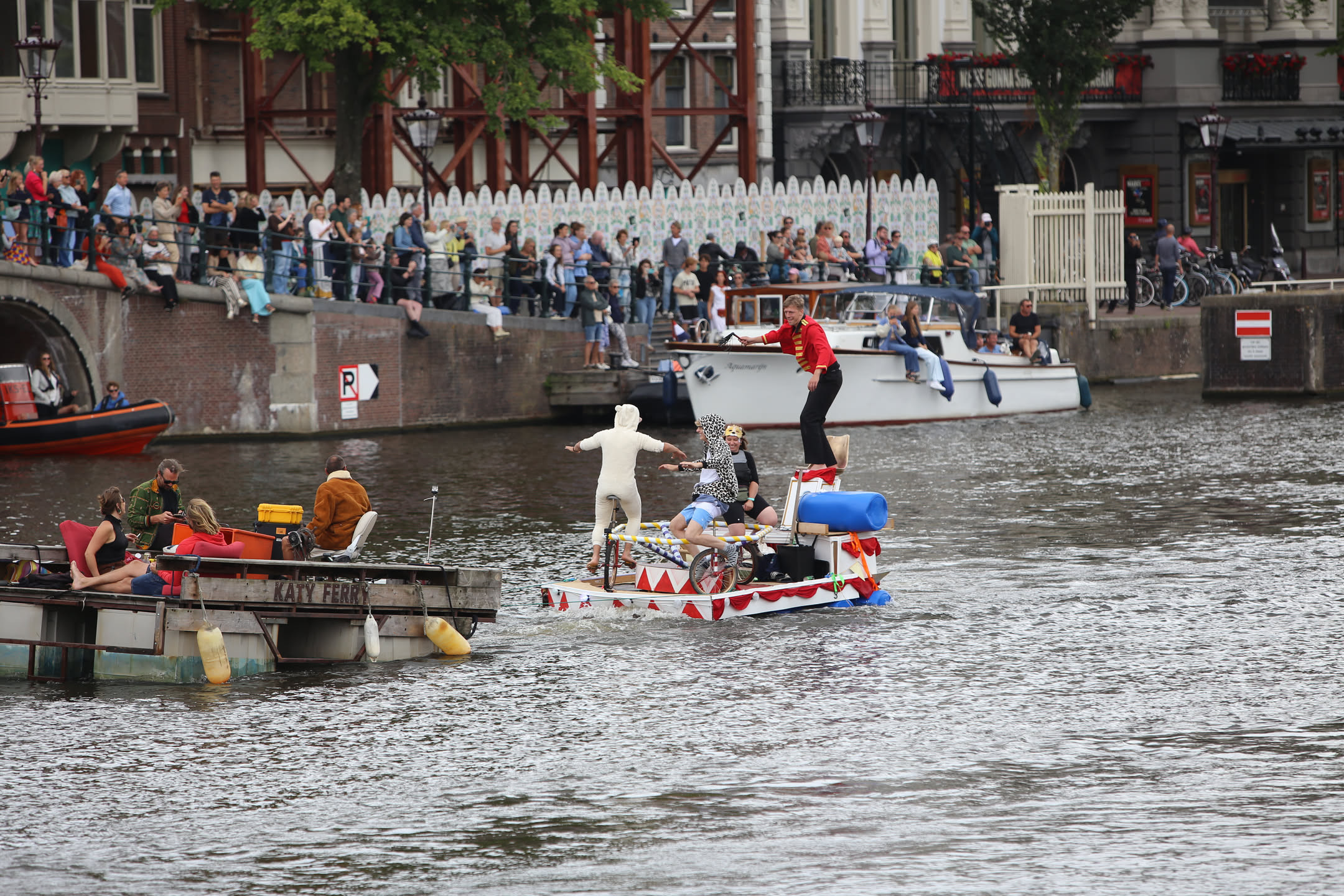 three people on a unicycle-go-round floating on a canal while a fourth person whips them. they are being towed by another ramshackle barge labeled "Katty Ferry"