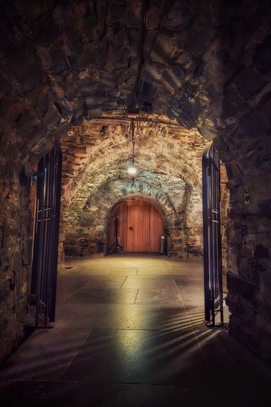 A low-angle shot shows a vaulted stone corridor underneath Christ Church Cathedral in Dublin. The corridor has rough-hewn stone walls and a matching ceiling, with a clean stone floor. Two black wrought iron gates are open in the foreground, inviting the viewer deeper into the scene. A large wooden door is visible at the end of the corridor, bathed in a warm light from a single bulb. The light casts long, dramatic shadows across the floor.