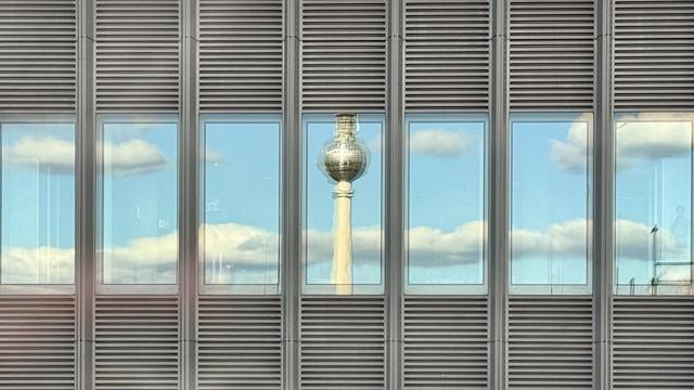 Narrow office building windows reflecting the blue sky and nice weather clouds. The middle one shows the top of the Berlin TV tower. It’s blurred, probably because of multiple window panes.