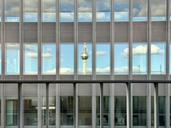 A wider shot of the same narrow office building windows reflecting the blue sky and nice weather clouds. The middle one shows the top of the Berlin TV tower. It’s blurred, probably because of multiple window panes.