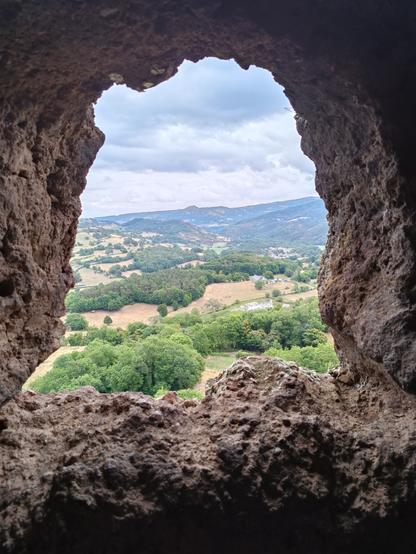 A travers une ouverture irrégulière dans une roche de couleur rose, vue sur un large paysage avec des bois, des prairies et des monts en forme de cônes. Le ciel est gris avec des nuages blancs et bleu foncé.