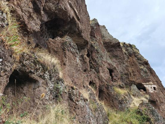 Vue en contre-plongée d'une paroi rocheuse de couleur rose foncé, avec des ouvertures à différents niveaux. De la végétation accrochée à la roche et un ciel gris-bleu nuageux.