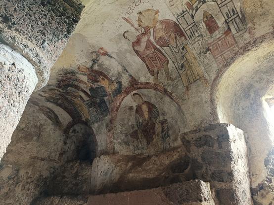 Vue en contre-plongée d'une partie des fresques de couleur rouge, jaune et noir, tracées sur les murs arrondis et la voûte d'une chapelle creusée dans la roche. On aperçoit le tombeau du Saint-Sépulcre, un soldat romain transperçant le flanc de Jésus, Marie dans un médaillon et la découverte du tombeau vide après la résurrection du Christ.
La lumière rentre par 2 ouvertures de chaque côté de ces fresques.