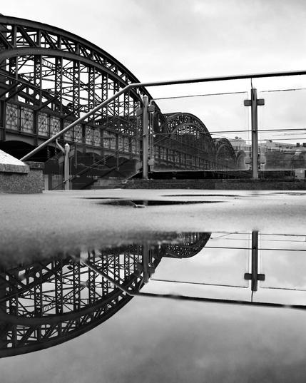 Das Bild zeigt eine stählerne Brückenkonstruktion, die aus mehreren großen Bögen und einem komplexen Geflecht aus Stahlträgern besteht, die ihr ein industrielles Erscheinungsbild verleihen. Im Vordergrund verläuft ein Geländer, das den Blick auf die Brücke teilweise verdeckt. Auf dem Boden befindet sich eine große Pfütze, in der sich sowohl die Brücke als auch das Geländer deutlich spiegeln. Der Himmel ist vollständig von grauen Wolken bedeckt.