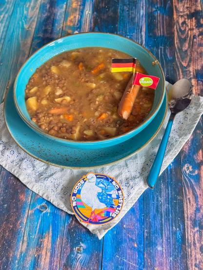 Das Bild zeigt eine Schüssel voller Linsensuppe mit Kartoffeln und Karotten, die in einem blauen Teller serviert wird. Im Teller steckt ein kleines deutsches Flaggenstäbchen und ein veganes Flaggenstäbchen, was darauf hinweist, dass die Suppe vegan ist. Neben der Schüssel liegt ein lila Gabel auf einem weißen Tuch mit Blumenmuster. Vor der Schüssel befindet sich ein kleiner, farbenfroher Aufkleber mit der Aufschrift "NO GODS NO MASTERS" und einem stilisierten Vogel. Die Schüssel steht auf einem blauen Holztisch, der mit einem abgenutzten Look versehen ist.