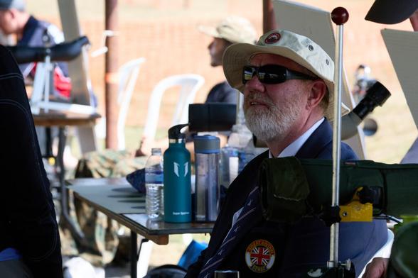 A suited man is seated. He wears a sun hat, sunglasses and a blue blazer with a badge embroidered with the Union Flag and the words "Great Britain Dewar".