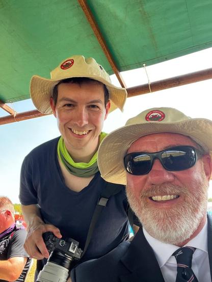 Two smiling men pose for a selfie. One is suited, wearing a white shirt and a tie bearing an emblem of crossed rifles. The other is wearing a thing blue t-shirt for the hot weather, has a green cooling towel around his neck and is carrying a Canon camera with a large lens.