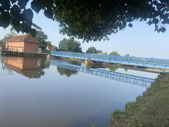 Ein blaues Wehr an einem blauen Fluss. Im Hintergrund ein Gebäude aus Ziegeln. Von oben hängen Zwrige ins Bild.