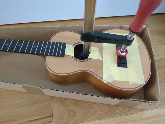 A ukulele with a big carpentry clamp through the sound hole, holding down the bridge. There are multiple layers of masking tape around the bridge to locate it.