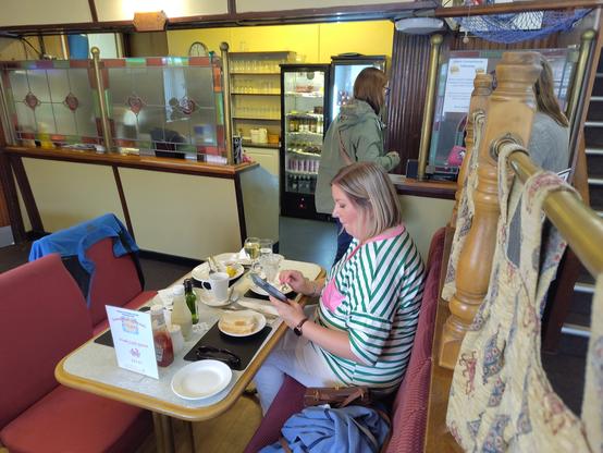 A photo of a traditional looking fish chip restaurant, with a lady at a table and a counter behind her. It seems as though this could have been taken 50 years ago or yesterday.
