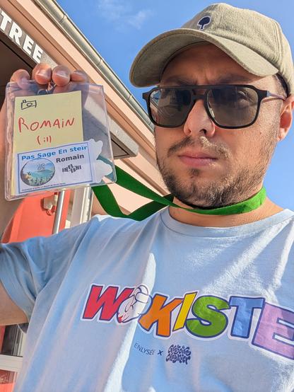 Selfie de moi avec une casquette, des lunettes de soleil, et un tee-shirt bleu clair avec écrit "wokiste", tenant mon badge pour le festival "Pas Sage En Steïr"