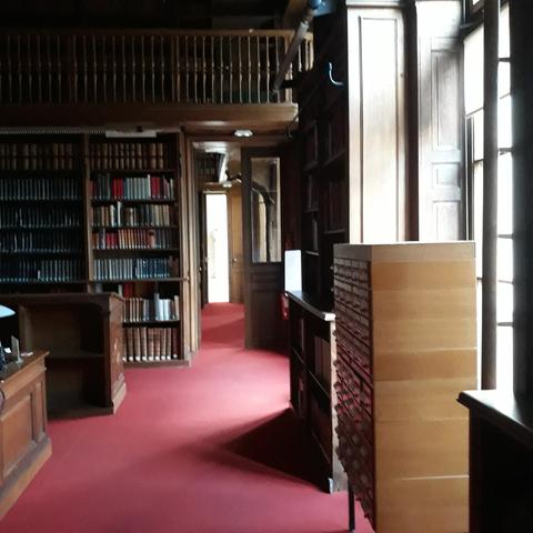 Vue de la salle d'accueil de la bibliothèque  de l'Arsenal, déserte : moquette rouge, étagères en bois pour les usuels, petite balustrade de la  coursive en mezzanine.