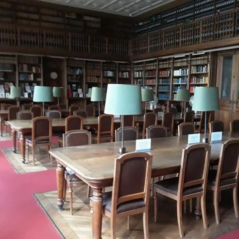 Vue de la salle de lecture avec ses tables en bois, ses chaises en bois et cuir, ses abats-jour verts. Les circulations autour de la salle sont en moquette rouge.